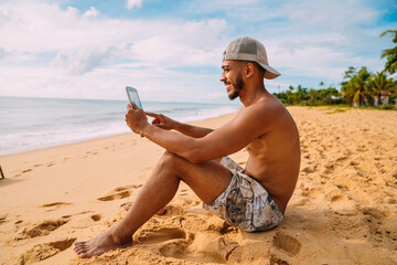 summer holidays, technology and internet concept. latin american man sitting on the sand, sunbathing on the beach with a tablet