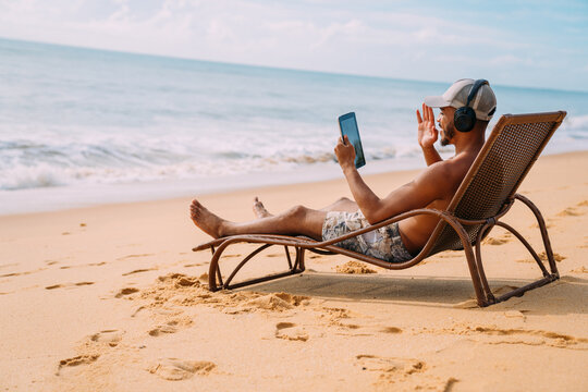 Man making a video call on summer vacation. latin american man sitting on the beach chair with headphones and a tablet