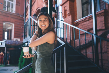 Fototapeta na wymiar Half length portrait of hipster girl with takeaway caffeine beverage and cellphone device smiling at camera during leisure in city, Asian female tourist with smartphone and coffee to go posing