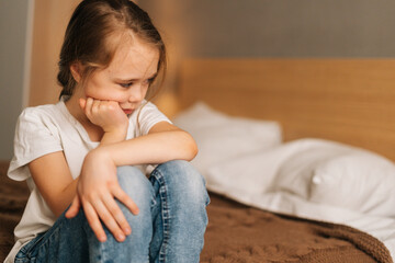 Close-up face of distraught beautiful little girl with wet eyes from tears sits on bed in bedroom and looking down. Depressed child feeling pressure, thinking of kid problems.