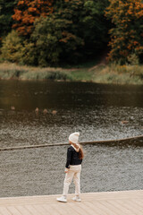 Fototapeta na wymiar little girl in a hat and black jacket walks near the lake in autumn