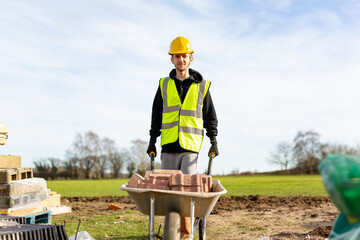 A young adult male builder wearing a high visibility vest and hard hat pushing a wheelbarrow full of bricks while on a building site