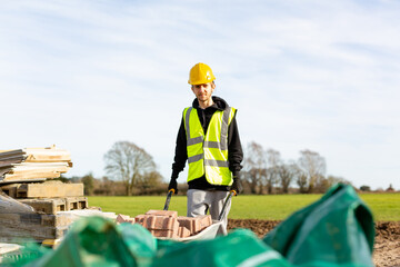 A young adult male builder wearing a high visibility vest and hard hat pushing a wheelbarrow full...