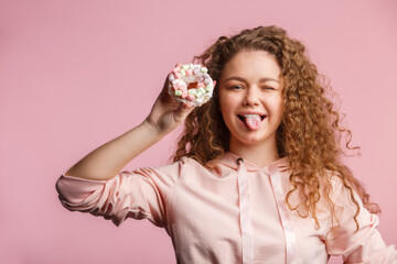 Young beautiful girl with curly hair holds a pink donut with marshmallows. Pink background.