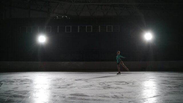 teen girl is preparing for competition of figure skating, junior female sportsperson is skating in dark stadium, moving against bright soffit