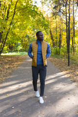 Fototapeta premium Positive african american young man in stylish clothes walks in late summer park on sunny warm warm day. Outdoor activity concept.