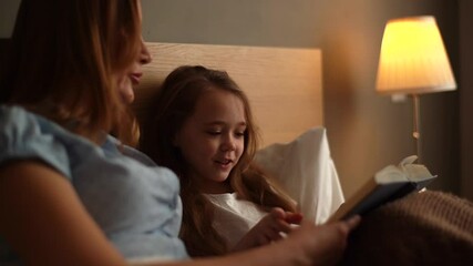Close-up portrait of happy young mother and adorable daughter reading together children book before going to sleep while lying in bed in dark nursery bedroom, near lamp. Tracking shot in slow motion. - Powered by Adobe