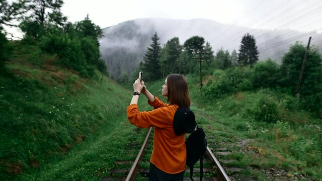 Woman hiker stands in the evening on the railway track in the mountains and makes photos and videos on a smartphone camera on a background of foggy forest. Morning walk in the mountains at dawn.