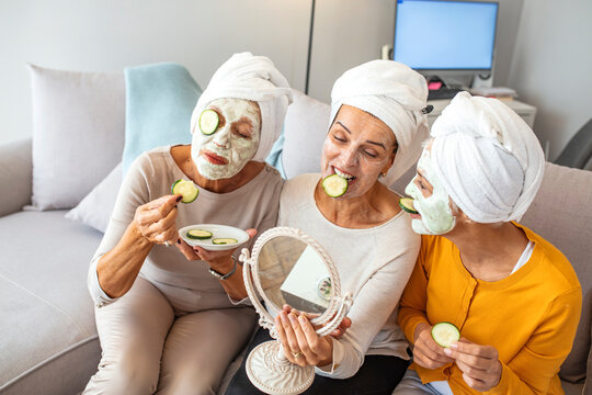 Diverse friends smiling while applying skincare face masks to their faces. Happy mature females laughing together sharing leisure time. Senior  friends having beauty treatment at home