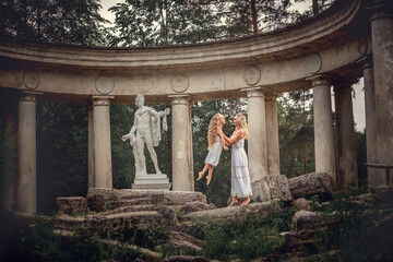 Beautiful blonde long-haired mother and daughter walking and dancing like two ancient goddess in the ruins of Colonnade Of Apollo in Pavlovsk park, Russia. Image with selective focus and toning