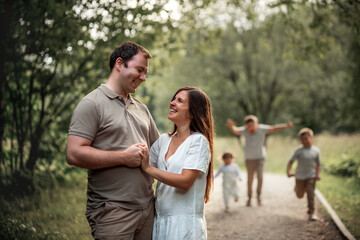 Fototapeta premium Happy married couple is hugging in the park, while their three kids are running towards them. Image with selective focus and toning.