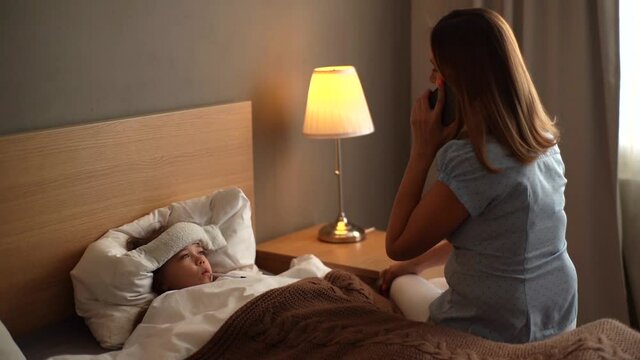 Worried young mother sitting on bed beside her sick daughter with high fever and calling on phone to family doctor. Mom measures temperature using thermometer of sick child lying under blanket at home