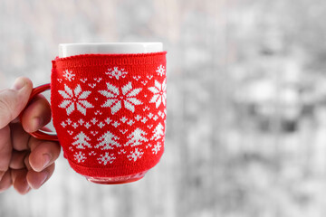 man's hand hold full Red cup with hot coffee or chocolate on background of beautiful snowy winter pine forest. Food and drink backdrop. Knitting ornament on red cup.  winter drink. No face.