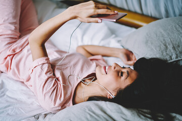 Obraz na płótnie Canvas Happy Asian woman in electronic headphones checking received email message while watching positive morning vlog, cheerful female blogger networking and listening music during lazy day in bedroom