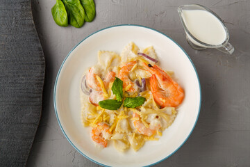 Farfalle pasta with shrimps in a creamy sauce on a gray plate on a concrete background next to a jug of cream.
