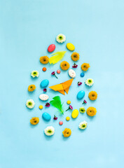 Creative Easter egg made of flowers, colored eggs, feathers, origami rabbits and butterflies, sweets on a blue background. 