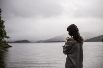 A beautiful shot of a Caucasian woman from behind wearing a cardigan in front of a lake
