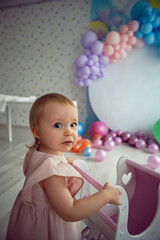 Fototapeta na wymiar baby girl in a pink dress carries a wooden stroller toy on a background of colorful balloons and a white round background
