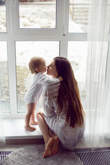 mother in a white robe sits with a child a blonde daughter at a large window of the house