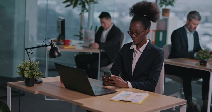 African american beautiful young woman sitting at office desk using phone application social media coworking with multi-ethnic staff at contemporary company.