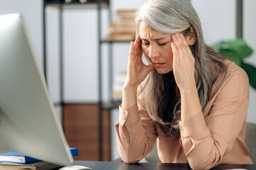 Tense tired senior gray-haired Asian, manager, business woman, freelancer thinks about a project or deal, massages her head with her hands, is stressed, she has a headache, sits at a table, need rest