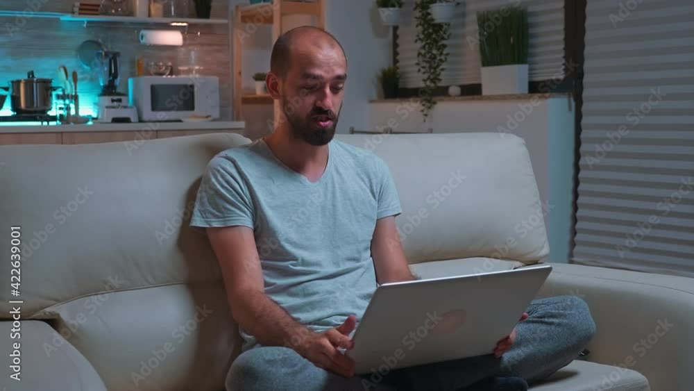 Poster focused man sitting on sofa in pajamas while talking with collegue during videocall conference using
