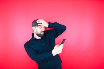 A bearded man in a black shirt holds a phone in his hand, holding his head. The bearded man is in shock