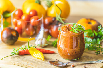 Homemade DIY natural canned hot plum sauce chutney with chilli or tkemali in glass jar standing on wooden table with ingredients.