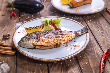 Grilled fish on a plate with vegetables, bell pepper, coarse salt on a brown wooden background.