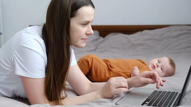 Young mother using laptop studying online working from home in internet lying on bed near cute baby infant, smiling focused mom typing on computer surfing web looking at screen enjoying distant job