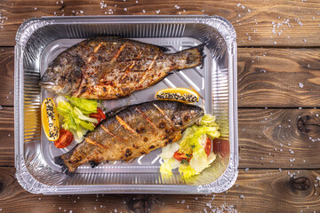 Grilled fish with vegetables, bell pepper, coarse salt, laid in an aluminum container on a brown wooden background.