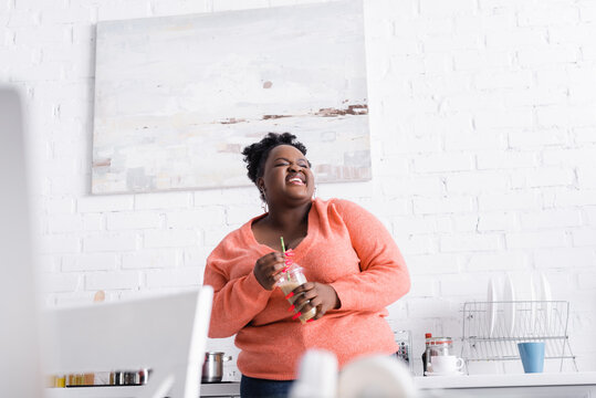 Cheerful African American Plus Size Woman Holding Plastic Cup With Tasty Smoothie In Kitchen