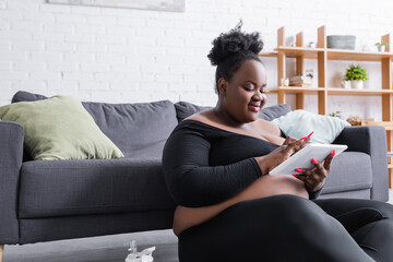 pleased african american plus size woman in sportswear using digital tablet in living room