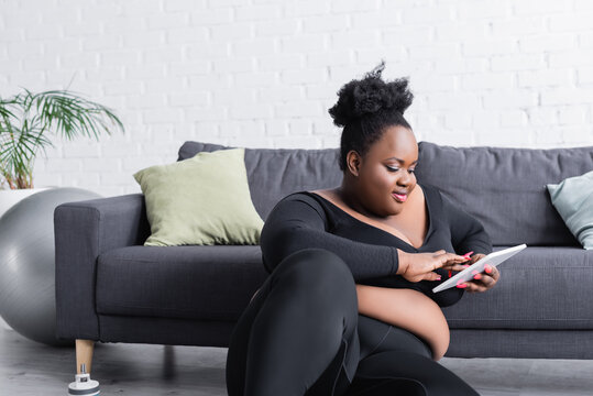 happy african american plus size woman in sportswear using digital tablet in living room - Powered by Adobe