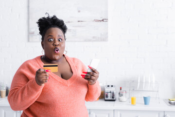 shocked african american plus size woman holding credit card and smartphone