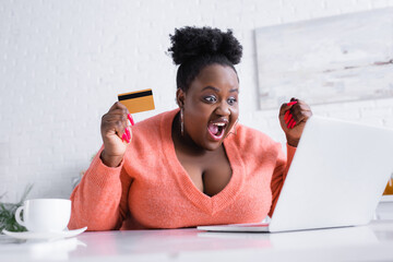 amazed african american plus size woman holding credit card and looking at laptop