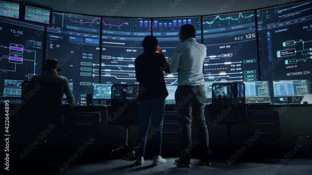 Poster In the System Control Room Project Manager and IT Technical Engineer Have Discussion, they're surrounded by Multiple Monitors with Graphics. Big Monitor Shows Interactive Server Blockchain Info.