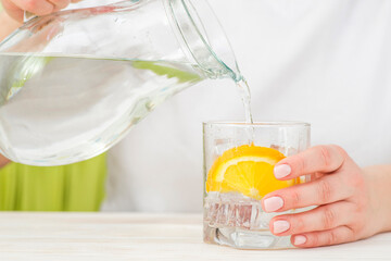 Female hands pouring water from the decanter into a glass beaker with lemon and ice. Health and...