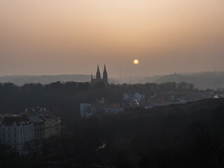 Aerial view over Prague 2 district from Nusle bridge, nuselsky most with typical architecture, old defense wall of Vysehrad castle and Basilica of St. Peter and Paul towers with orange sun in haze