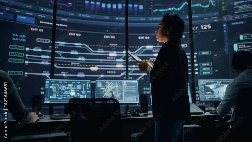 Poster Young Multiethnic Female Government Employee Uses Tablet Computer in System Control Monitoring Center. In the Background Her Coworkers at Their Workspaces with Many Displays Showing Technical Data.