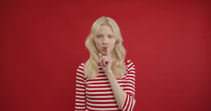 Stop talking. Dissatisfied strict young blonde woman making quiet gesture, shushing with finger on lips, asking for silence or secrecy. Indoor studio shot isolated on red background