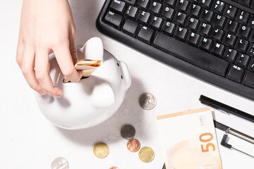 Child puts money into the piggy bank on white background with clipboard, coins and keyboard, long banner format. Financial education for children concept.