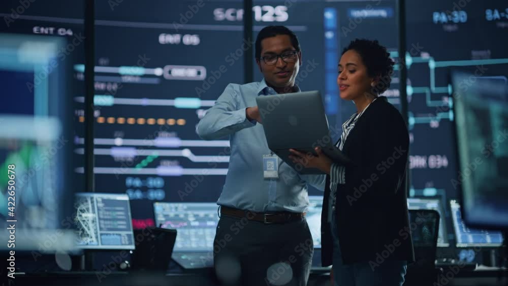 Poster In the System Control Room Project Manager and IT Technical Engineer with Laptop Have Discussion, surrounded by Multiple Monitors with Graphics. Big Monitor Shows Interactive Server Blockchain Info.