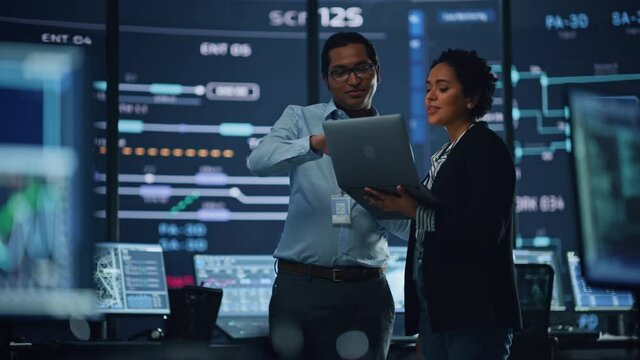 In the System Control Room Project Manager and IT Technical Engineer with Laptop Have Discussion, surrounded by Multiple Monitors with Graphics. Big Monitor Shows Interactive Server Blockchain Info.