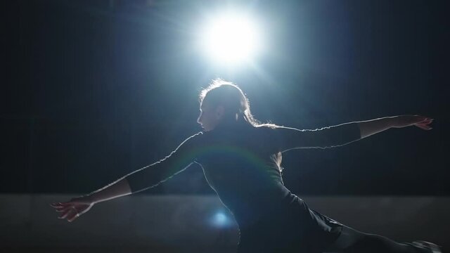 young female figure skater is training alone in dark ice rink, jumping and spinning, talented professional sportswoman