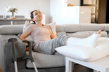 Obraz premium Young woman with pain expression on couch at home with orthopedic plaster massaging leg. Fracture of the leg or foot. Concept of rehabilitation and healing physiotherapy. Orthopedics and Traumatology.