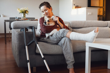 Adult woman in her late twenties on couch at home with crutches and orthopedic plaster caress the...