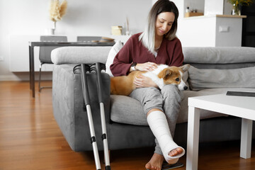 Adult woman in her late twenties on couch at home with crutches and orthopedic plaster caress the...