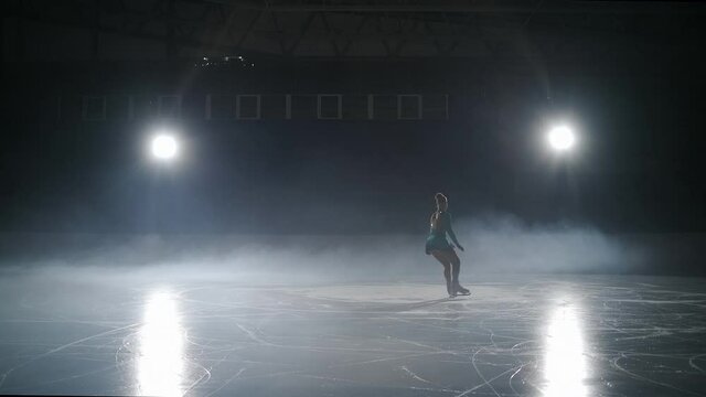 young female figure skater is performing on ice rink in darkness, small figure is moving against bright soffit inside dramatic fog