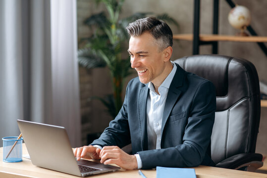 Influential successful Caucasian adult businessman or manager in a formal suit uses a laptop, communicates via video conferencing, online meetings, sits office, types on a keyboard, smiles friendly